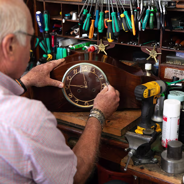 איש מבוגר במעבדה מכוונן שעון שולחן עץ בסביבת כלים ומכשור טכני.