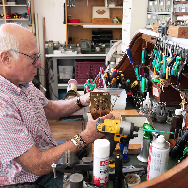 גבר מבוגר עם משקפיים ואביזרי יד בודק מנגנון שעון עליו שולחן עבודה מלא בכלי עבודה ומברגות.