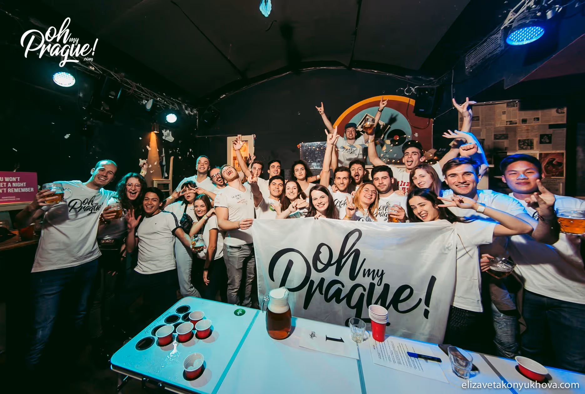 Group of young adults at a party holding a banner that reads 'Oh my Prague!' with drinks and red solo cups on a table in front.