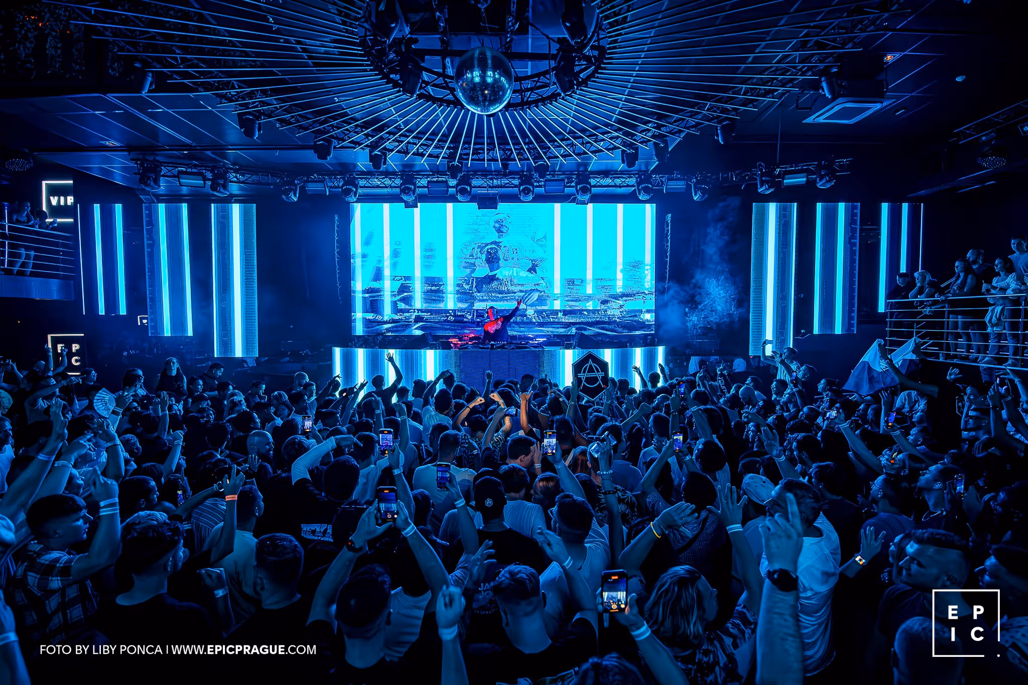 Crowd at a nightclub watching a DJ perform on a stage with bright blue LED lights.