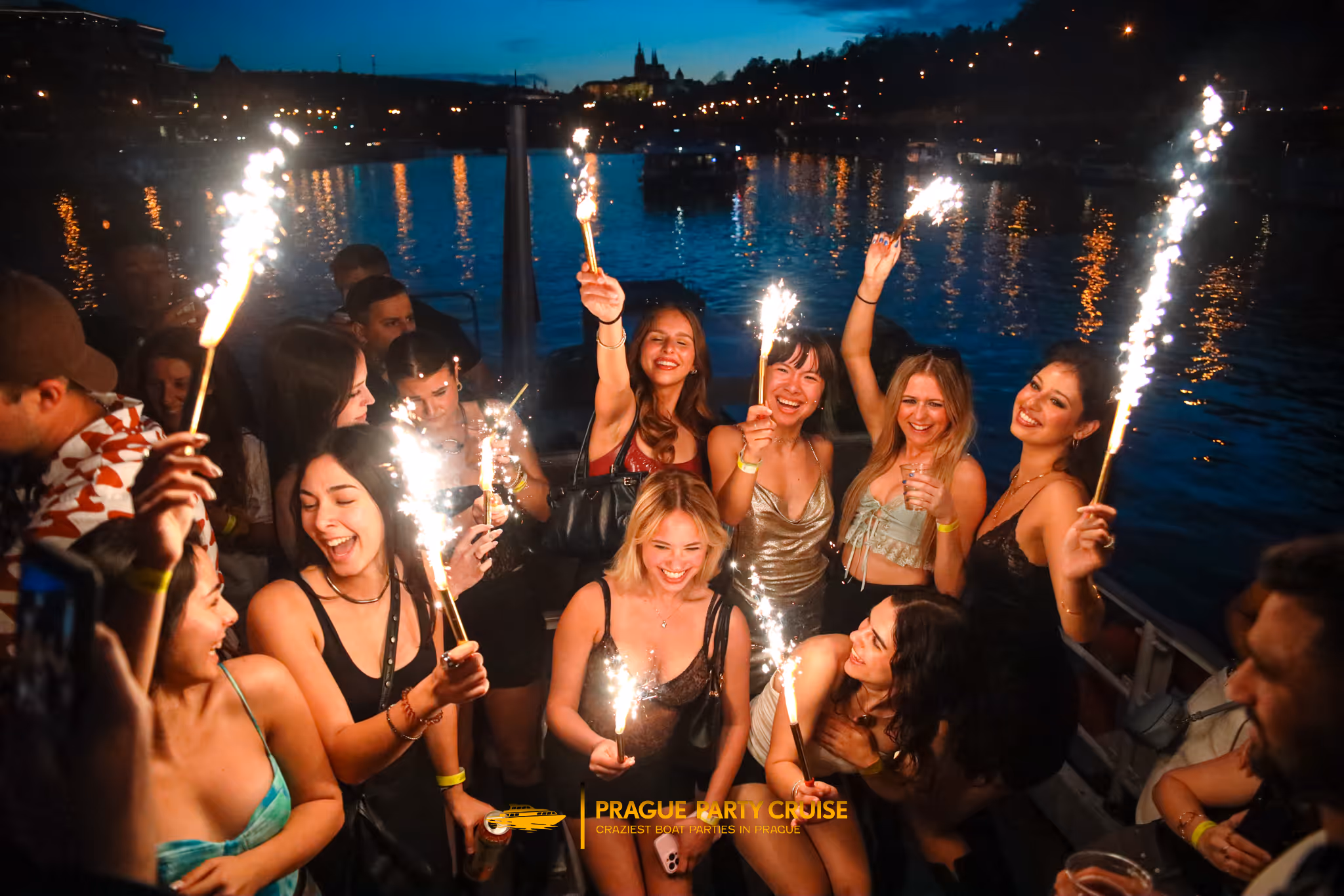 Group of young people celebrating on a boat at night holding sparklers with a city skyline and water in the background.