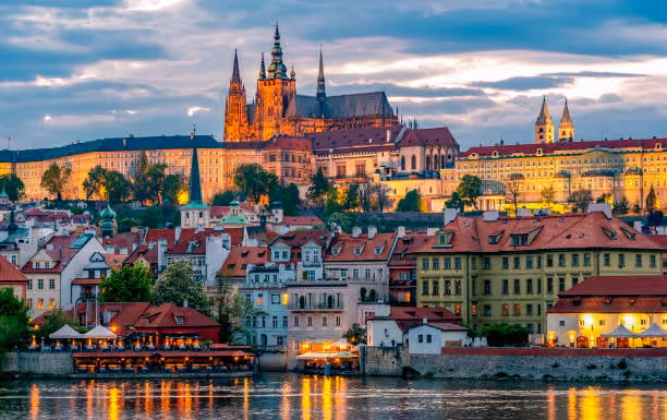 View of Prague Castle illuminated at dusk with historic buildings and the Vltava River in the foreground.