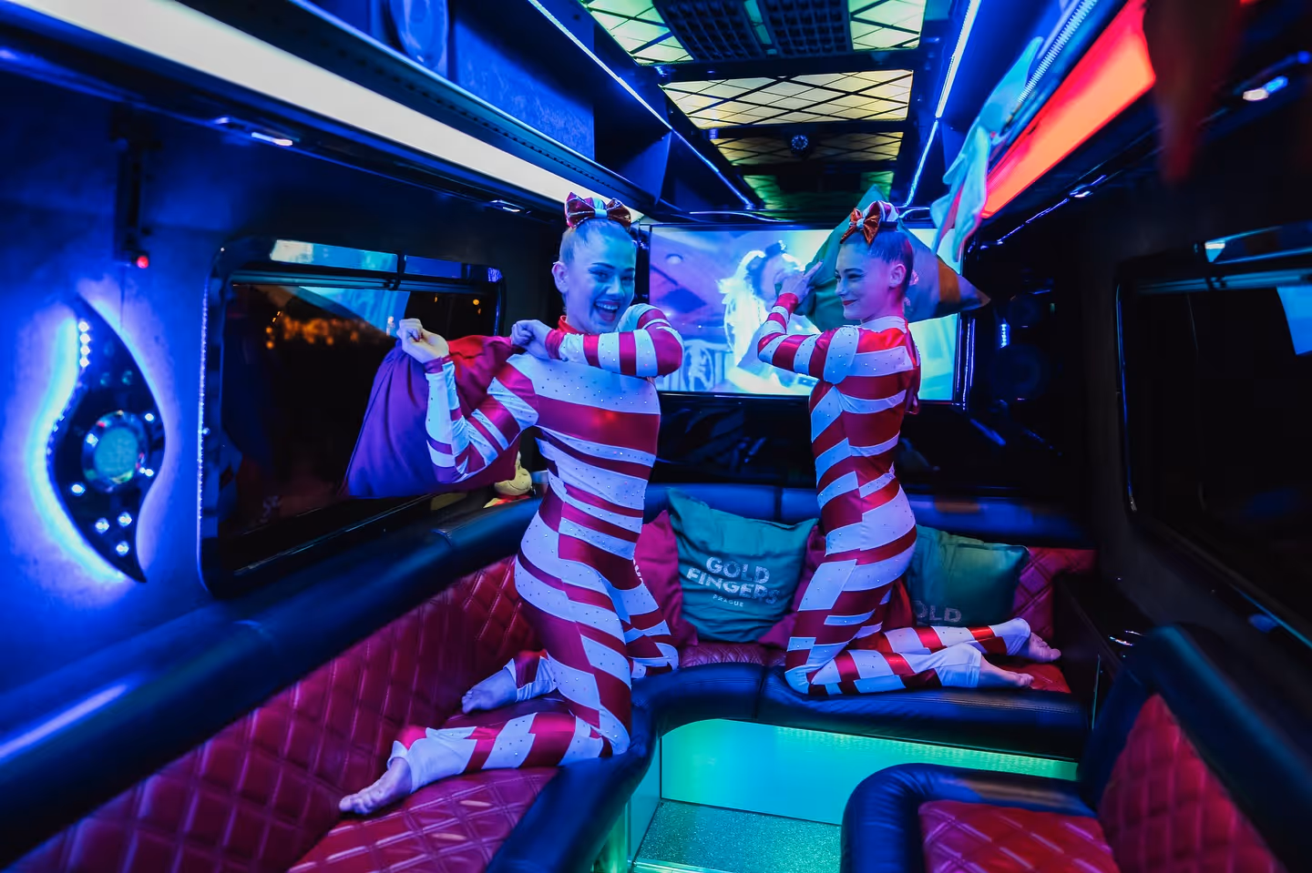 Two women in red and white striped costumes having a pillow fight inside a neon-lit party bus.