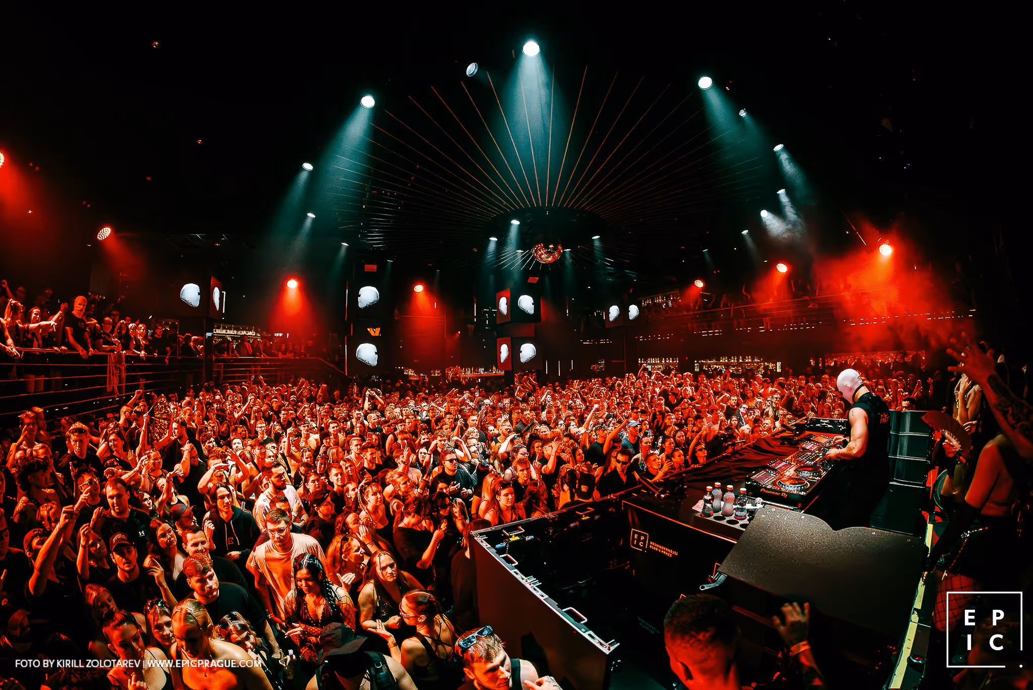 Crowded nightclub with DJ wearing a white mask performing on stage amid red and green lighting.