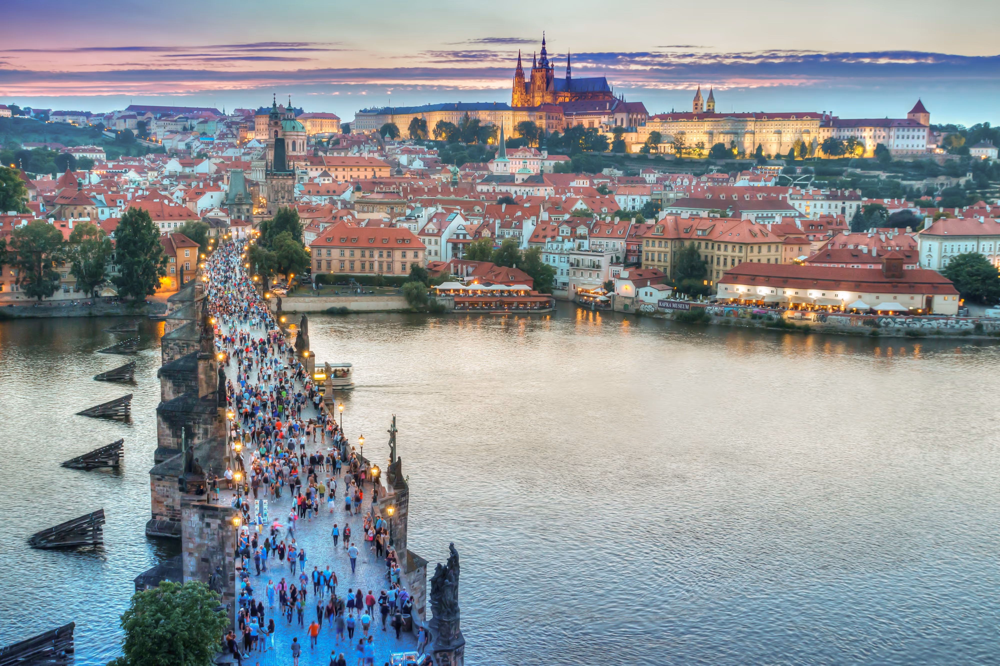 Crowded Charles Bridge over the Vltava River with Prague Castle illuminated in the background at sunset.