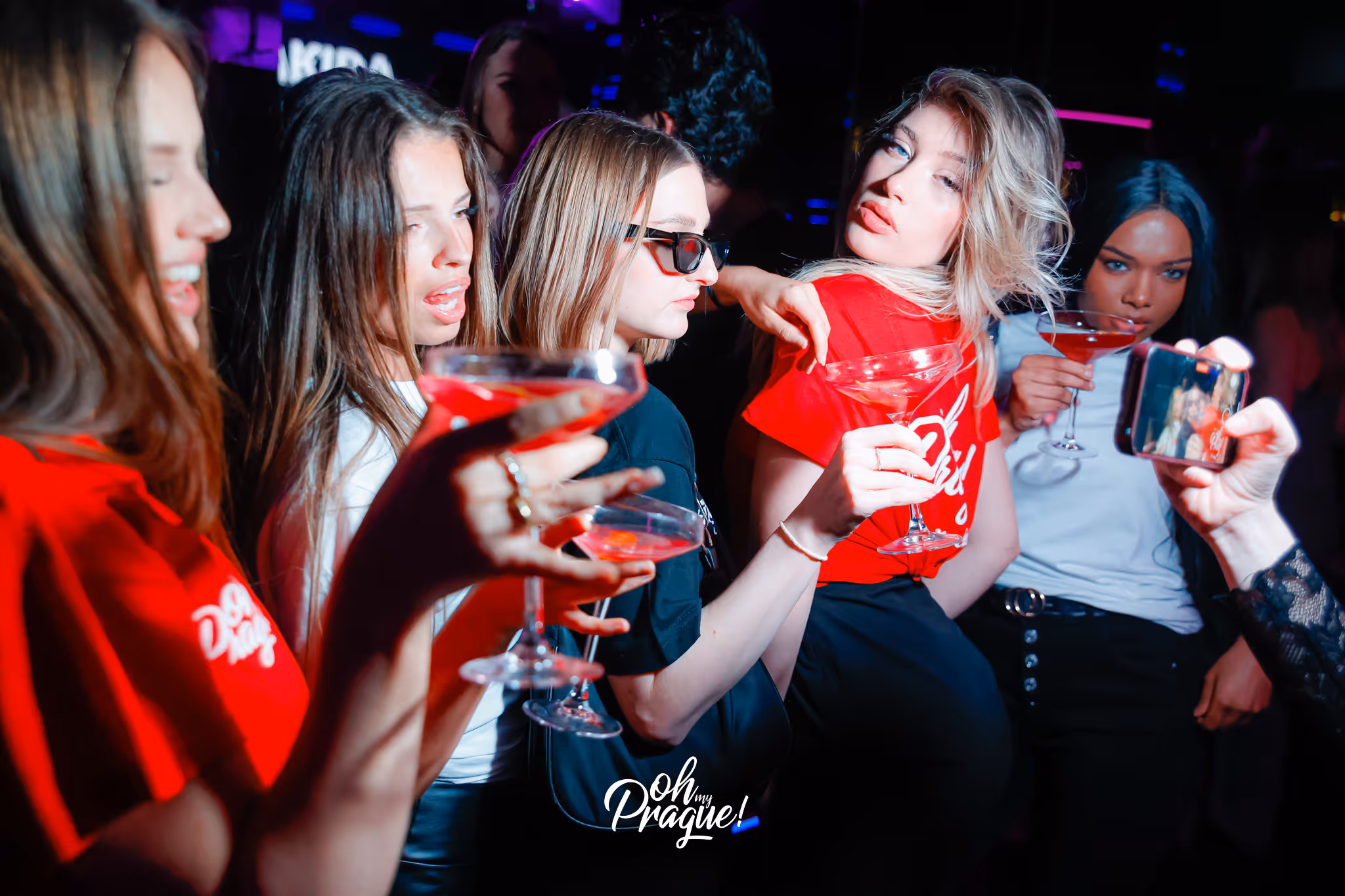 Group of five young women holding cocktails posing for a selfie in a dimly lit club.