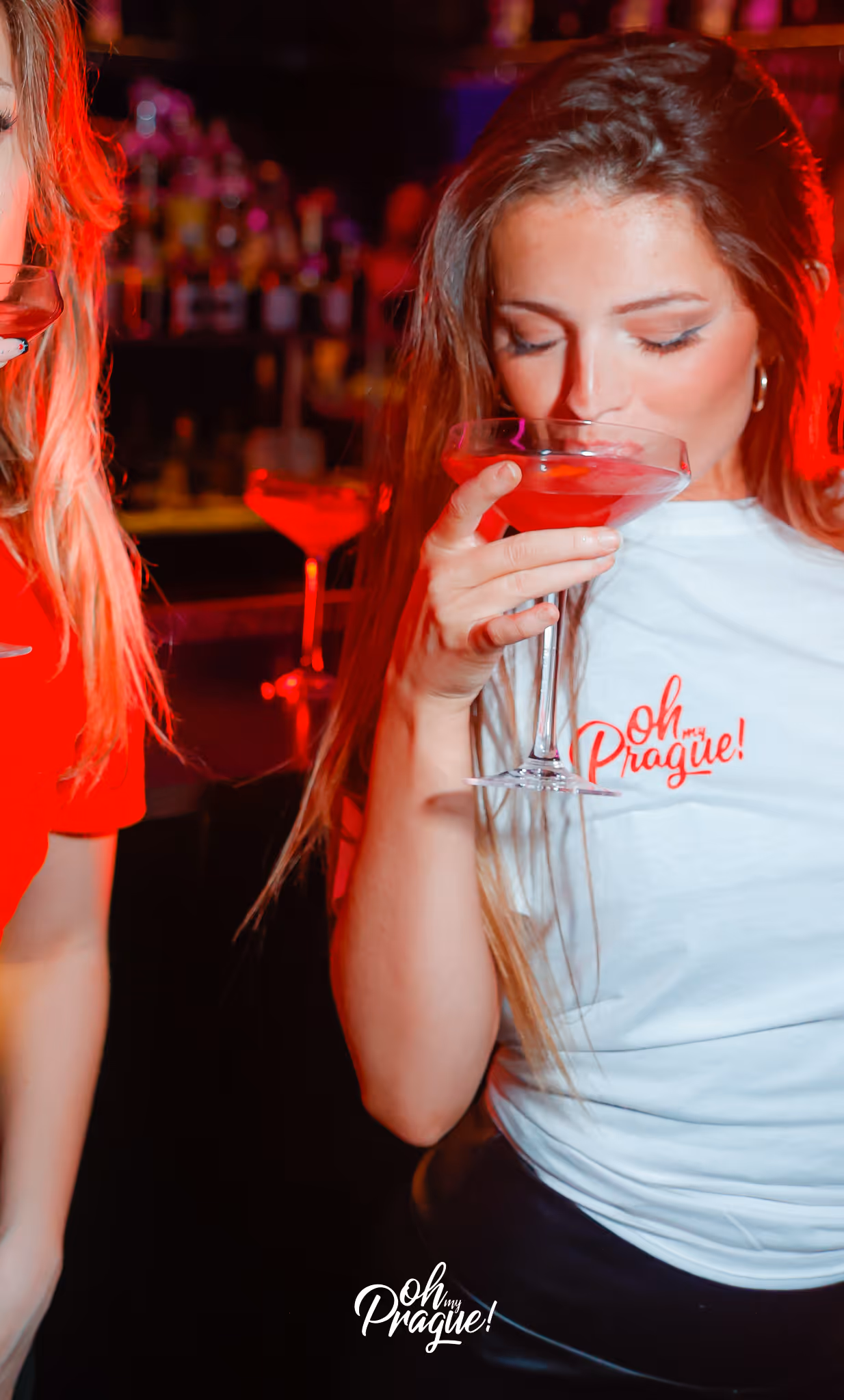 Young woman in a white 'Oh my Prague!' t-shirt sipping a red cocktail in a dimly lit bar.