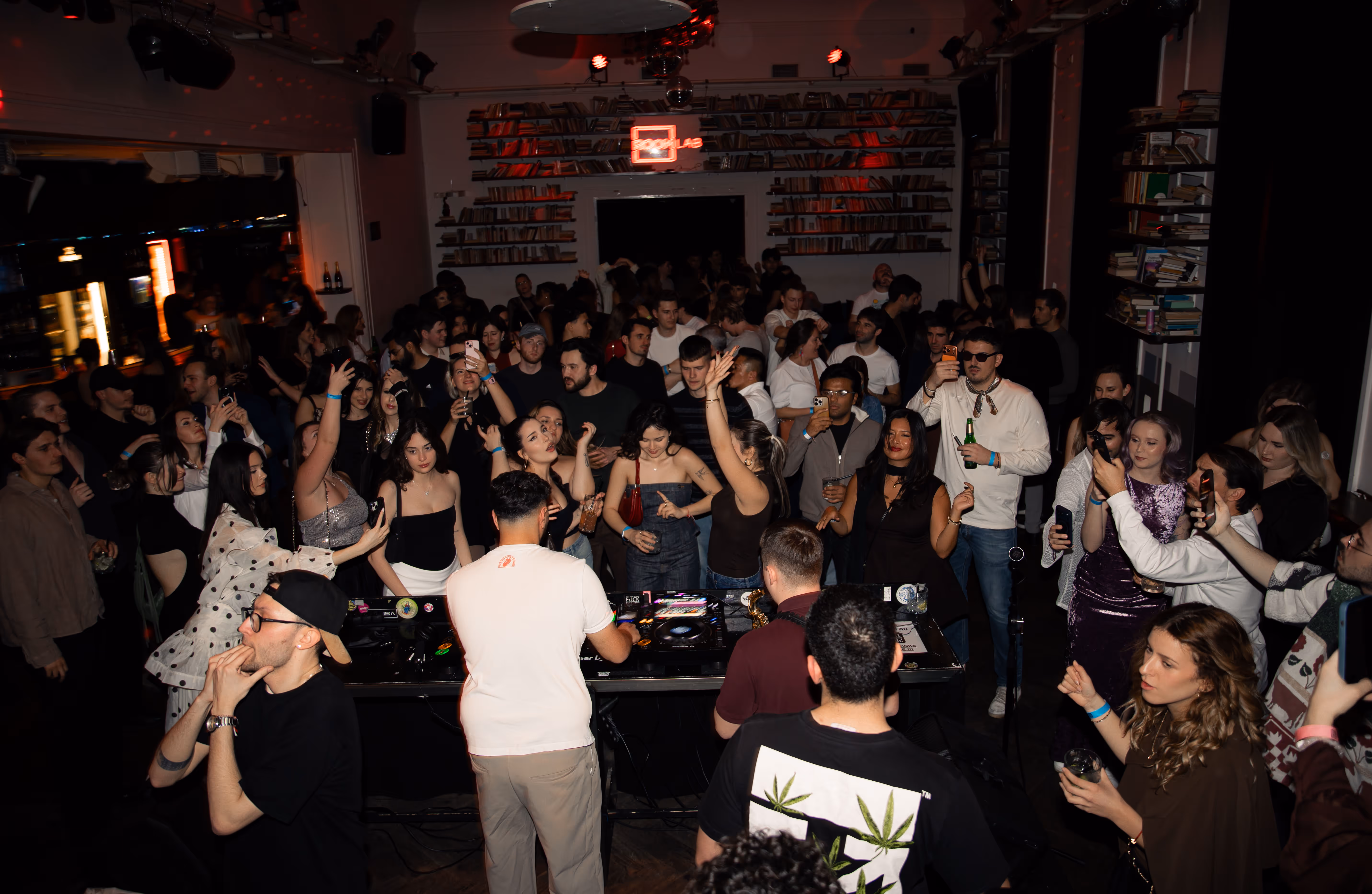 Crowd of people dancing and taking photos around a DJ performing in a dimly lit club with shelves of books on the walls.