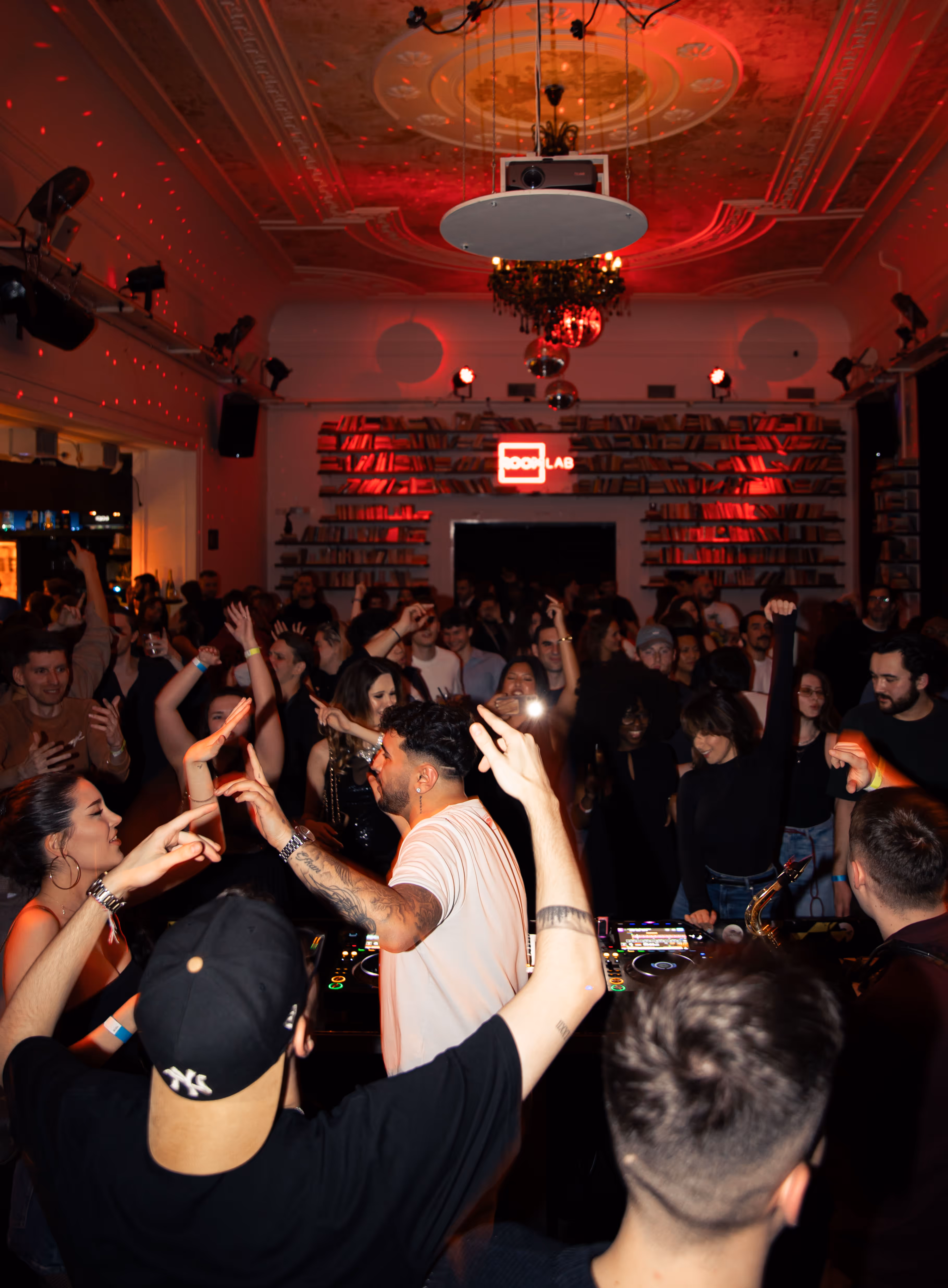 Crowded nightclub with people dancing and DJ playing music under red lighting and a bookshelf in the background.