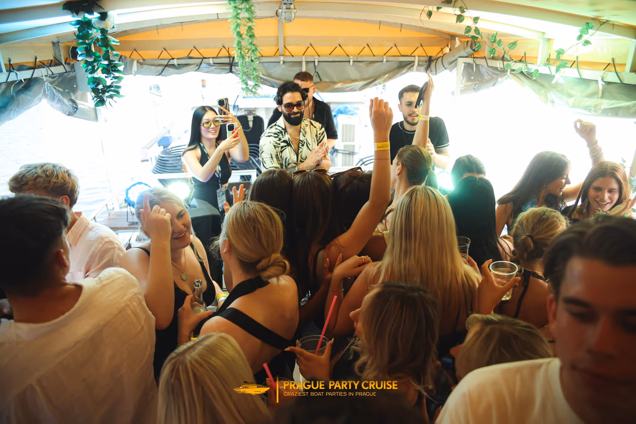 Crowd of people enjoying a lively party on a boat with a DJ playing music and attendees dancing and taking photos.