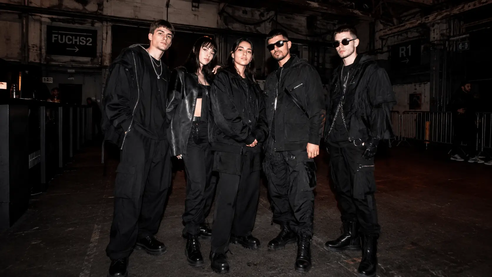 Prompt: A group of stylish young people wearing all-black, avant-garde streetwear and chunky boots, standing in an industrial warehouse setting. Underground techno fashion aesthetic, flash photography style, high contrast --ar 16:9