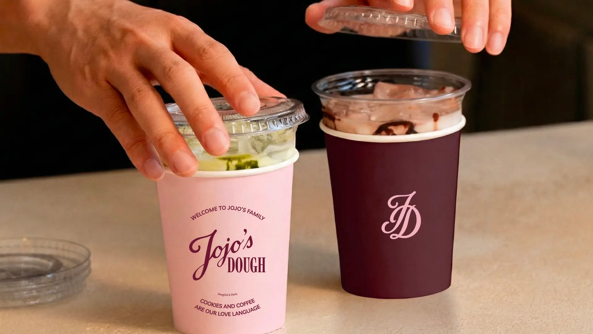 Deux gobelets à emporter Jojo’s Dough avec couvercles transparents, tenus en main sur un comptoir
