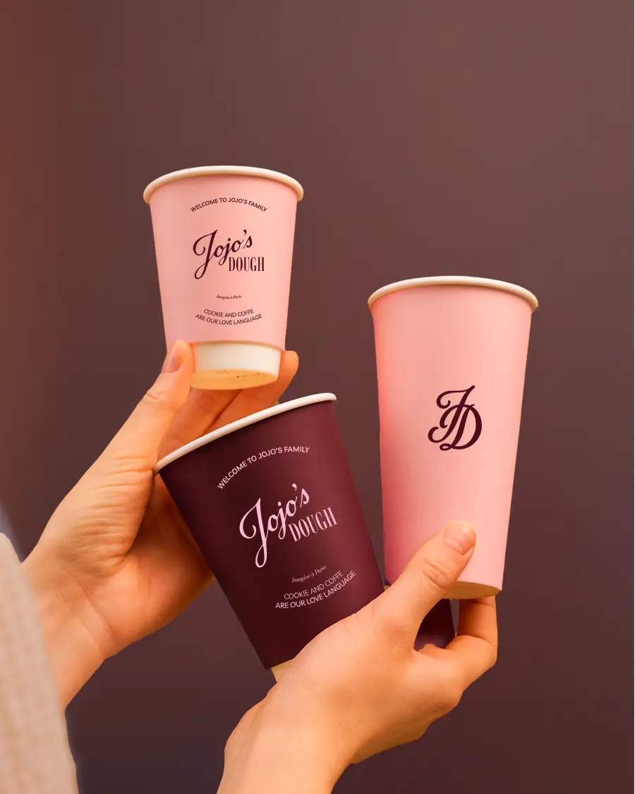 Trois gobelets à emporter Jojo’s Dough tenus en main, avec logos et couleurs de la marque