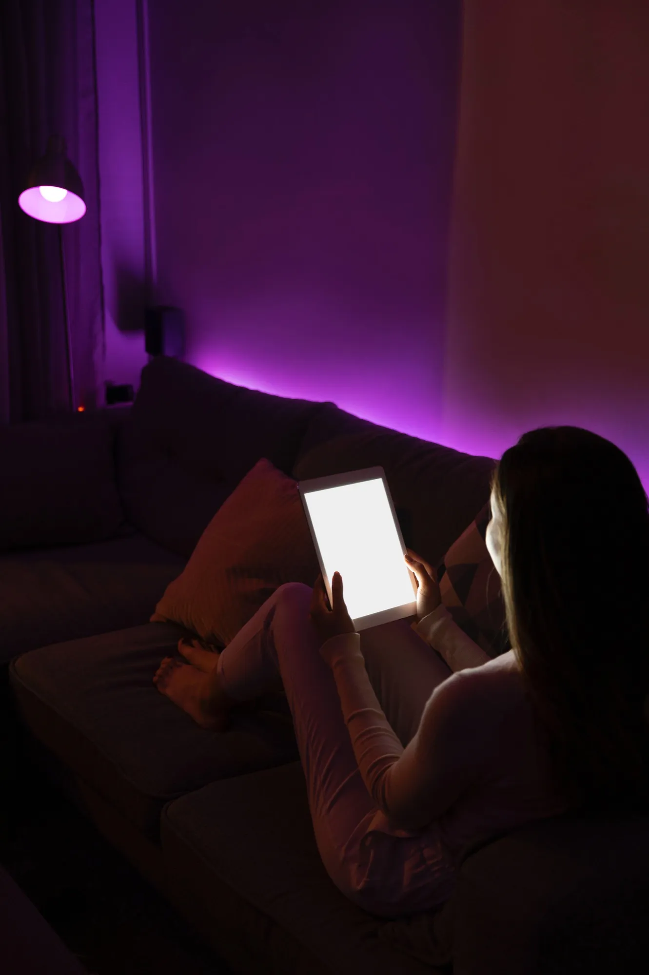 Person sitting on a couch in a dimly lit room with purple ambient lighting, holding and looking at a brightly lit tablet.