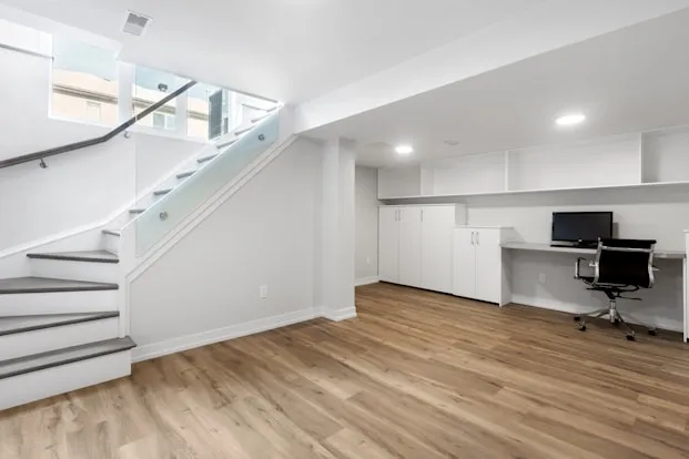 Modern basement room with wooden flooring, white built-in cabinets, a desk with a computer, and a staircase with glass railing.