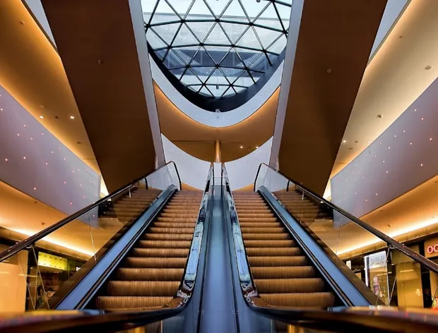 Upward view of two parallel escalators in a modern building with a geometric glass ceiling above.