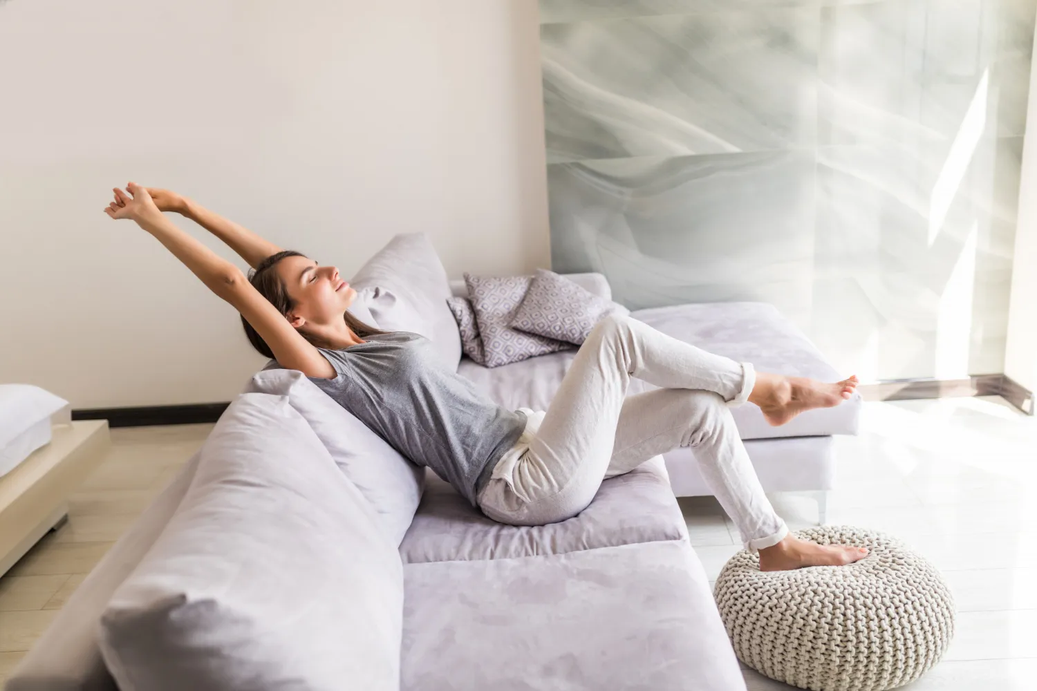 Woman stretching her arms back while relaxing on a light gray sofa with feet resting on a knitted pouf in a bright living room.