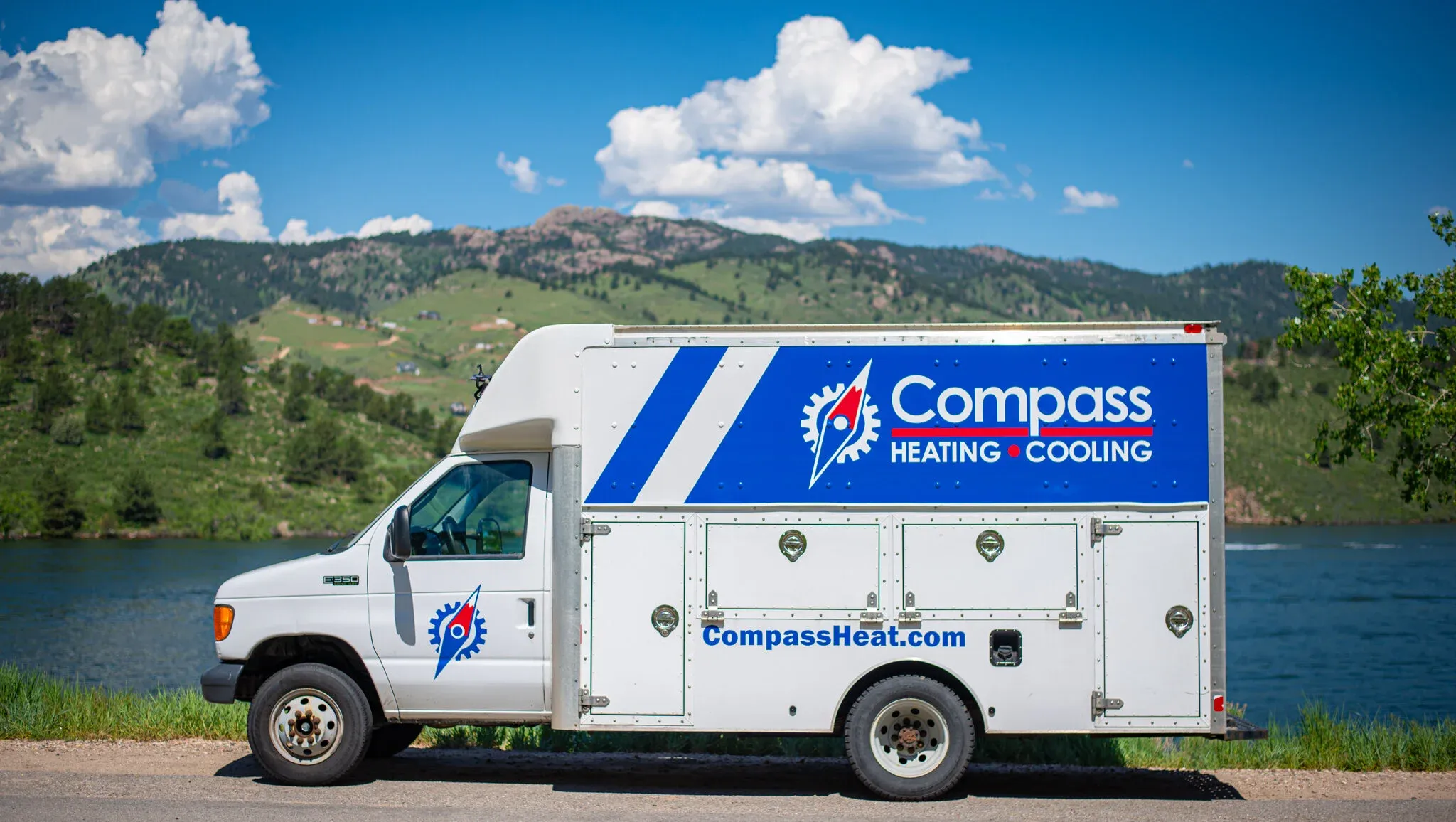 White Compass Heating and Cooling service truck parked beside a river with green hills and blue sky in the background.