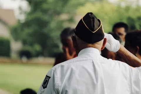 Veteran in white uniform and black cap saluting outdoors with blurred people and greenery in background.