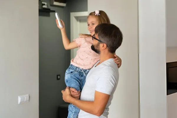 Smiling man holding a young girl who is holding up a remote control inside a home.