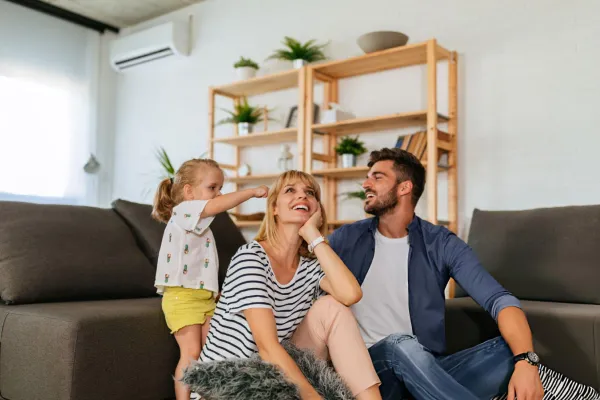 A smiling family of three sitting and playing together in a cozy living room with wooden shelves and plants in the background.