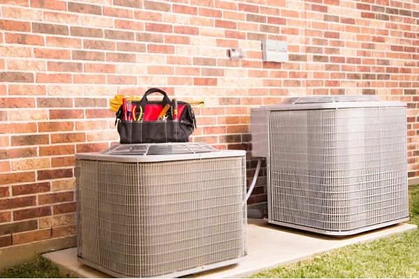 Two outdoor air conditioning units next to a brick wall with a black tool bag placed on top of one unit.