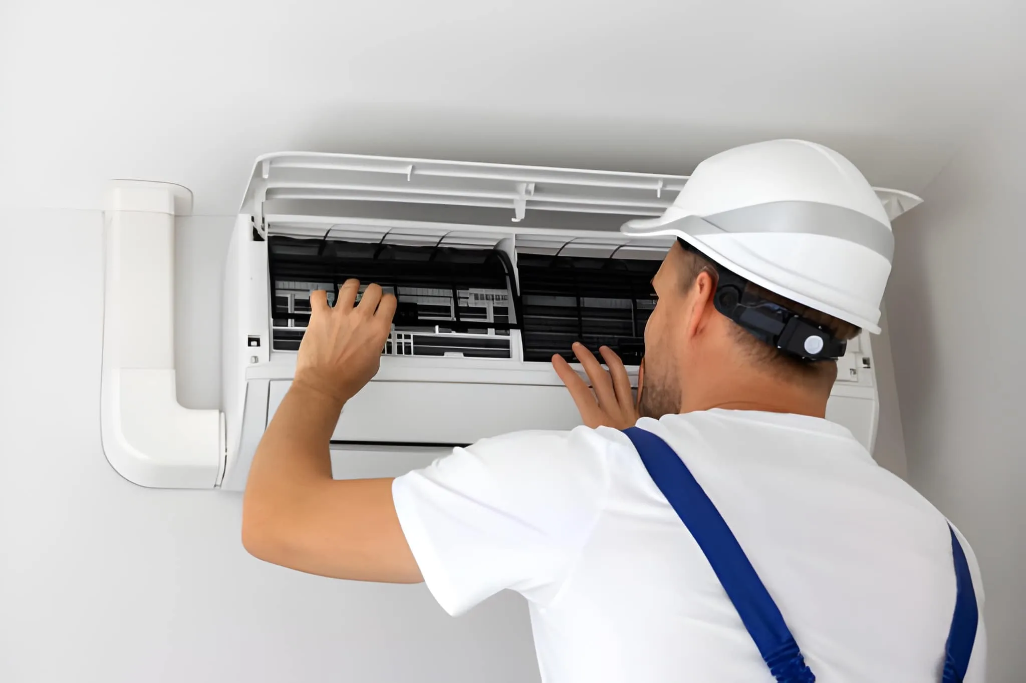Technician wearing a white hard hat and blue suspenders inspecting the filter of a wall-mounted air conditioner.