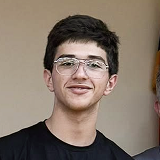 Young man with short dark hair and glasses smiling against a plain background.