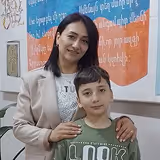 Smiling woman with her hand on the shoulder of a young boy standing in front of a colorful bulletin board with text.