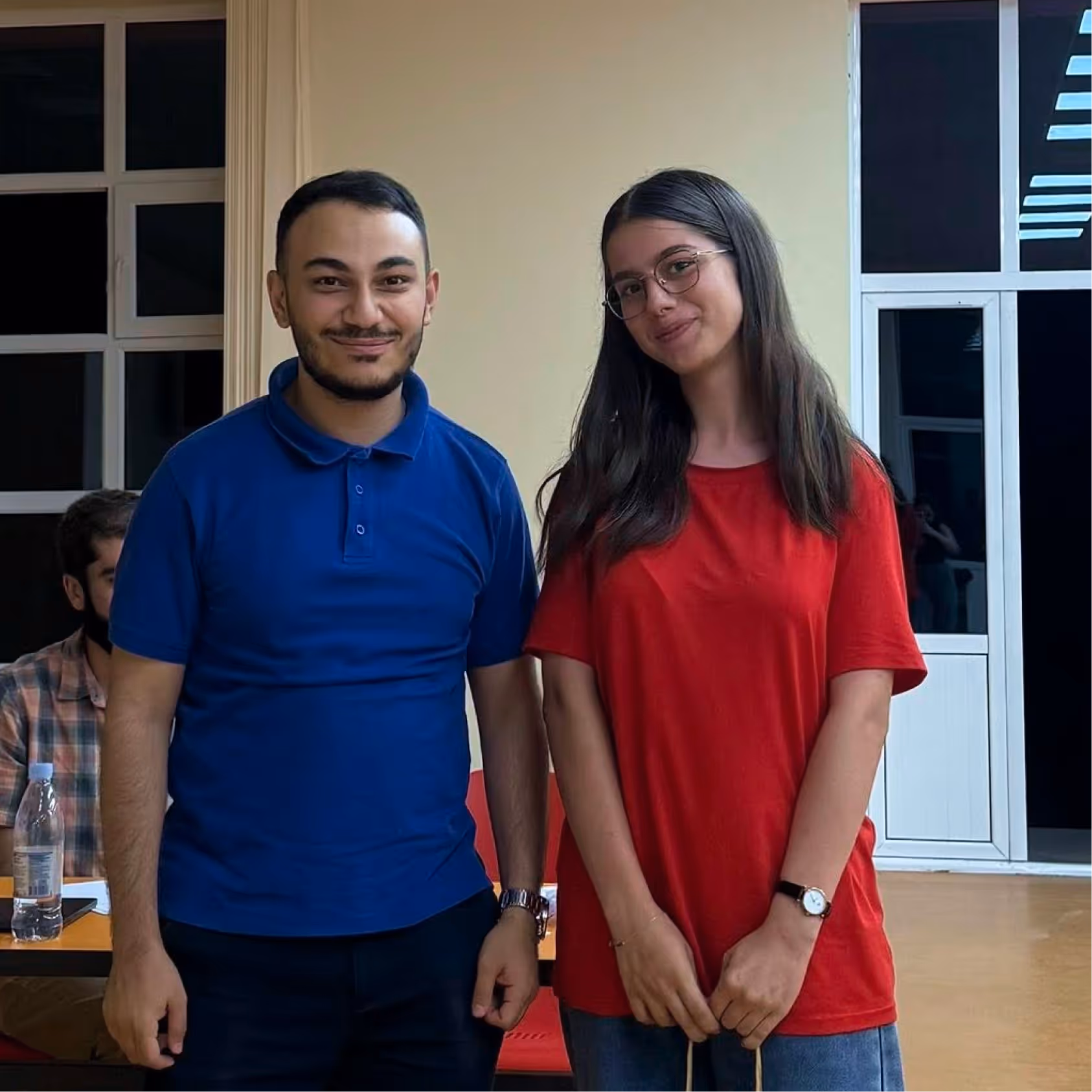 A man in a blue polo shirt and a woman in a red t-shirt smiling and standing indoors near windows.