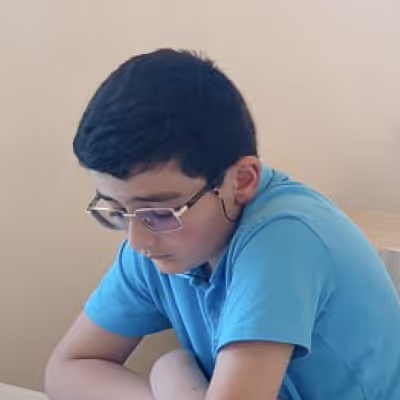 Boy with glasses in a blue shirt sitting at a table, looking downward with folded arms.