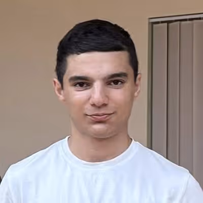 Portrait of a young man with short dark hair wearing a white t-shirt standing indoors near a window with vertical blinds.