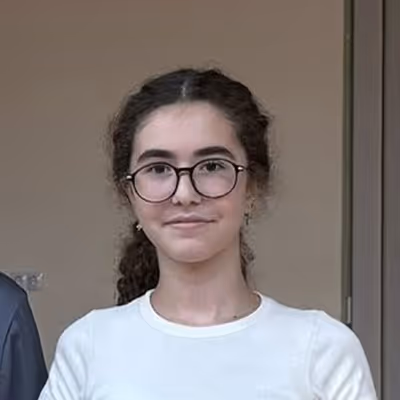 Young woman with curly hair pulled back, wearing black round glasses and a white top, posing indoors.