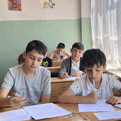 Four boys sitting at wooden desks in a classroom, focused on writing or reading papers under soft daylight from a window with sheer curtains.
