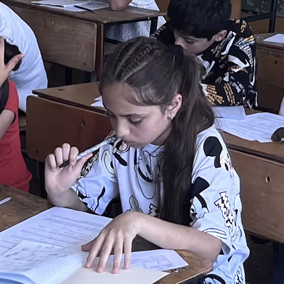 Young girl with braided hair sitting at a desk, holding a pen to her chin and reading a book attentively in a classroom.
