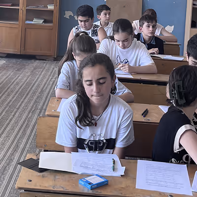 Students sitting at wooden desks in a classroom, focused on individual tasks with papers and pens in front of them.