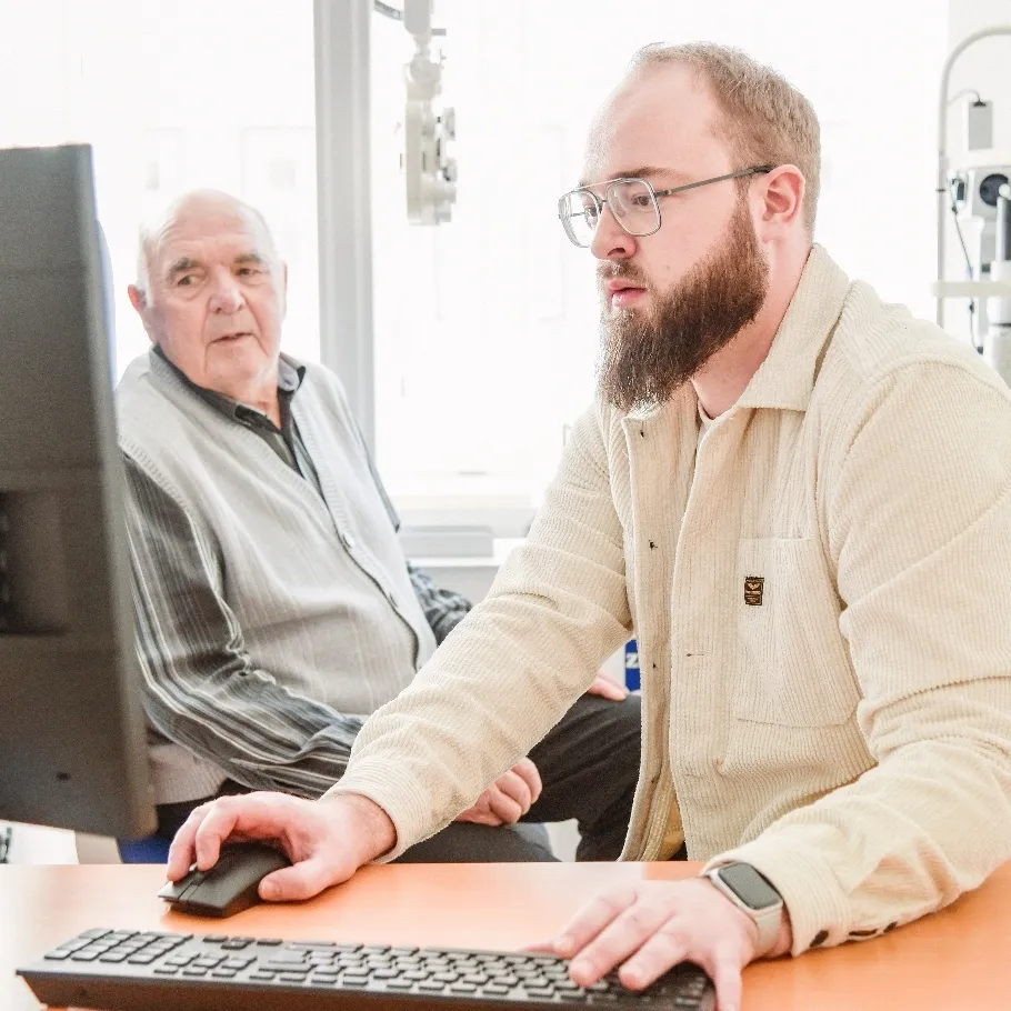 Ein junger Mann mit Brille und Bart benutzt eine Computermaus, während ein älterer Mann im Hintergrund auf einem Stuhl sitzt.