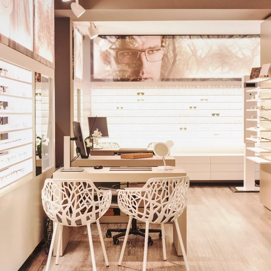 Modernes Optikergeschäft mit zwei weißen Designstühlen vor einem Beratungstisch, Brillen an den Wänden und großem Werbebild eines Mannes mit Brille.