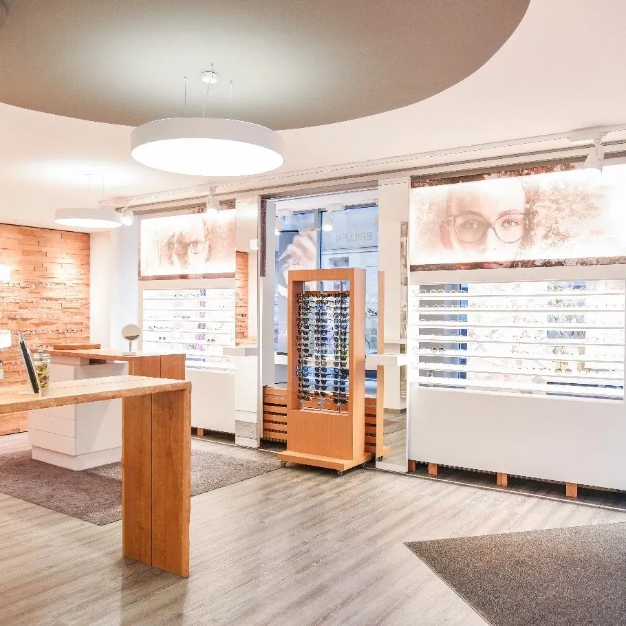 Helles, modernes Optikergeschäft mit Holztheken, Brillendisplays und großen Bildern von Menschen mit Brillen.