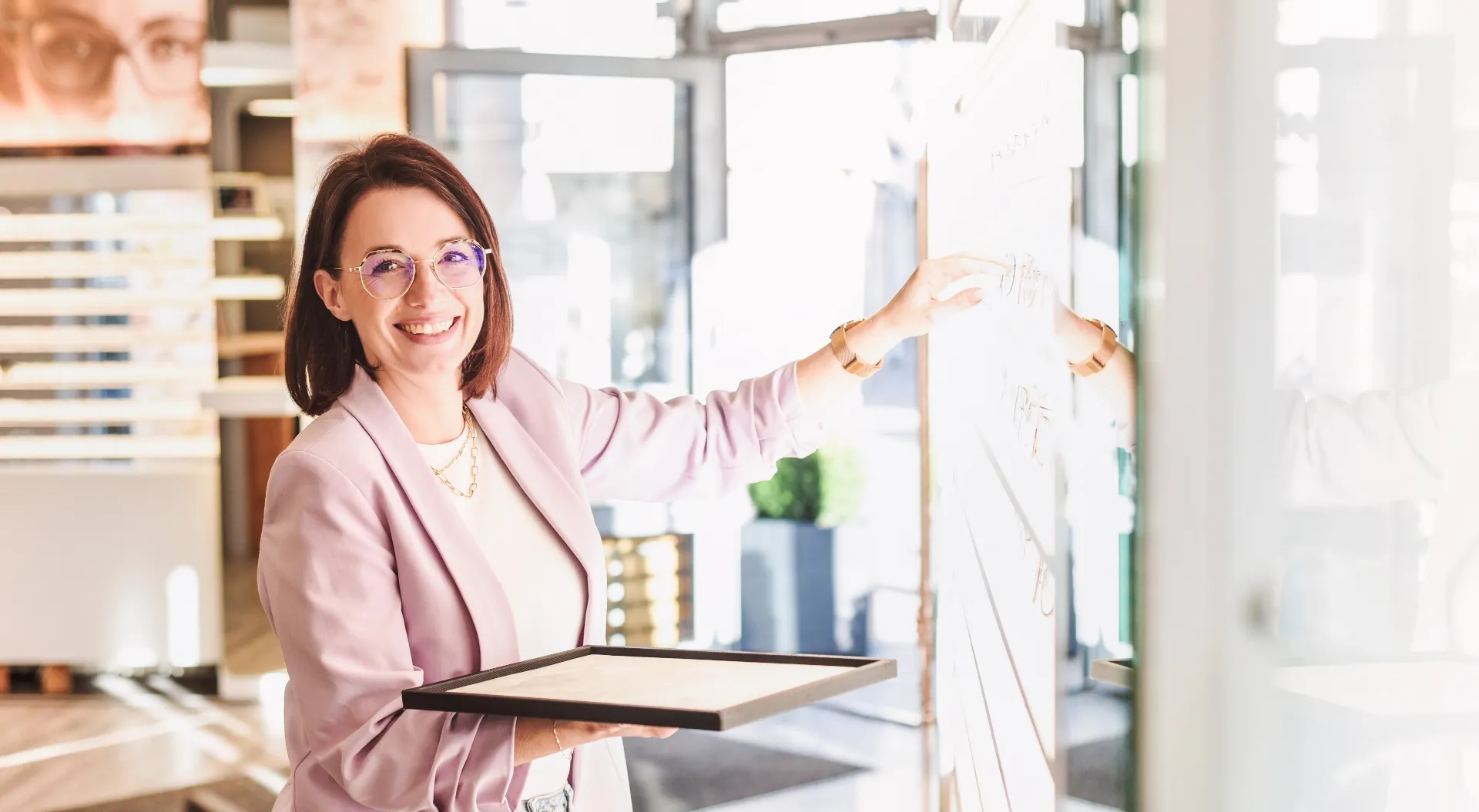 Lächelnde Frau mit Brille und rosa Blazer zeigt auf eine helle Tafel an der Wand und hält eine Ablage.
