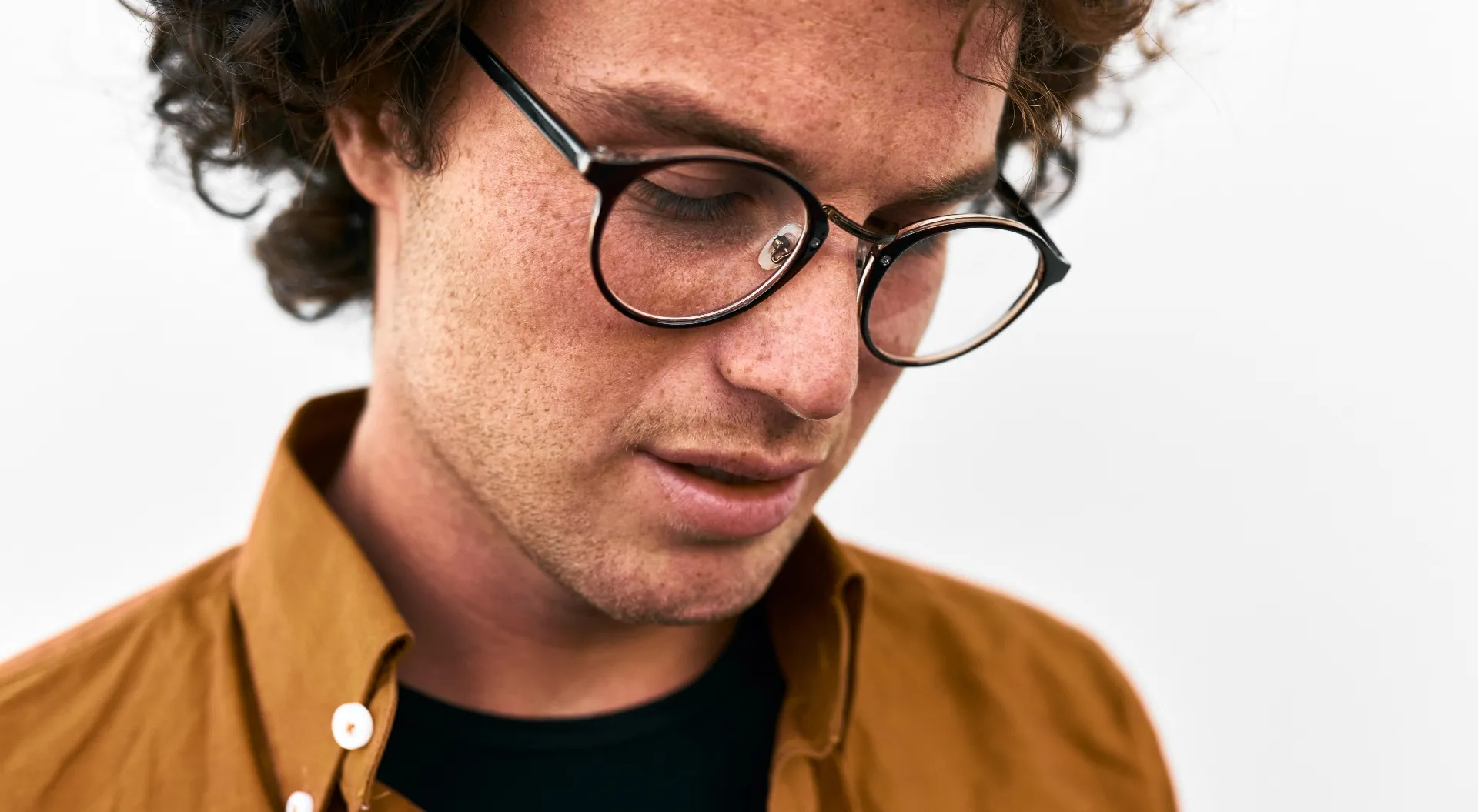 Nahaufnahme eines nach unten blickenden Mannes mit lockigem Haar und Brille, der ein braunes Hemd trägt.