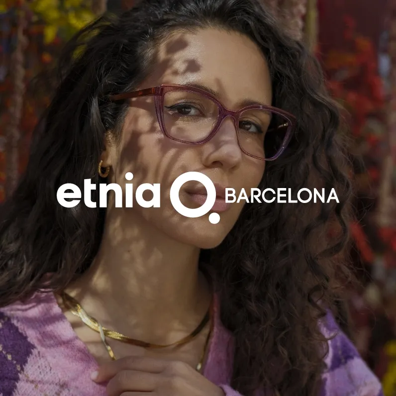 Markenbid Etnia Barcelona. Frau mit lockigem dunklem Haar und violetten Brillengestell vor herbstlichem, farbigem Hintergrund.