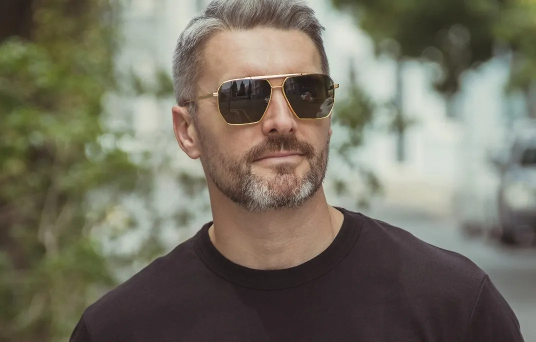 Mann mit grauem Bart und Haaren trägt goldgerahmte Sonnenbrille und schwarzes T-Shirt im Freien.