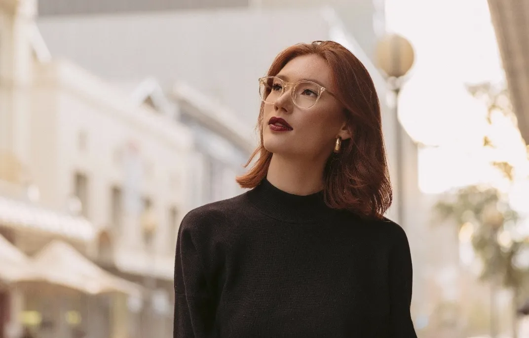 Frau mit roten Haaren, die eine schwarze Rollkragenpullover, transparente Brille und goldene Ohrringe trägt, schaut nachdenklich nach oben.