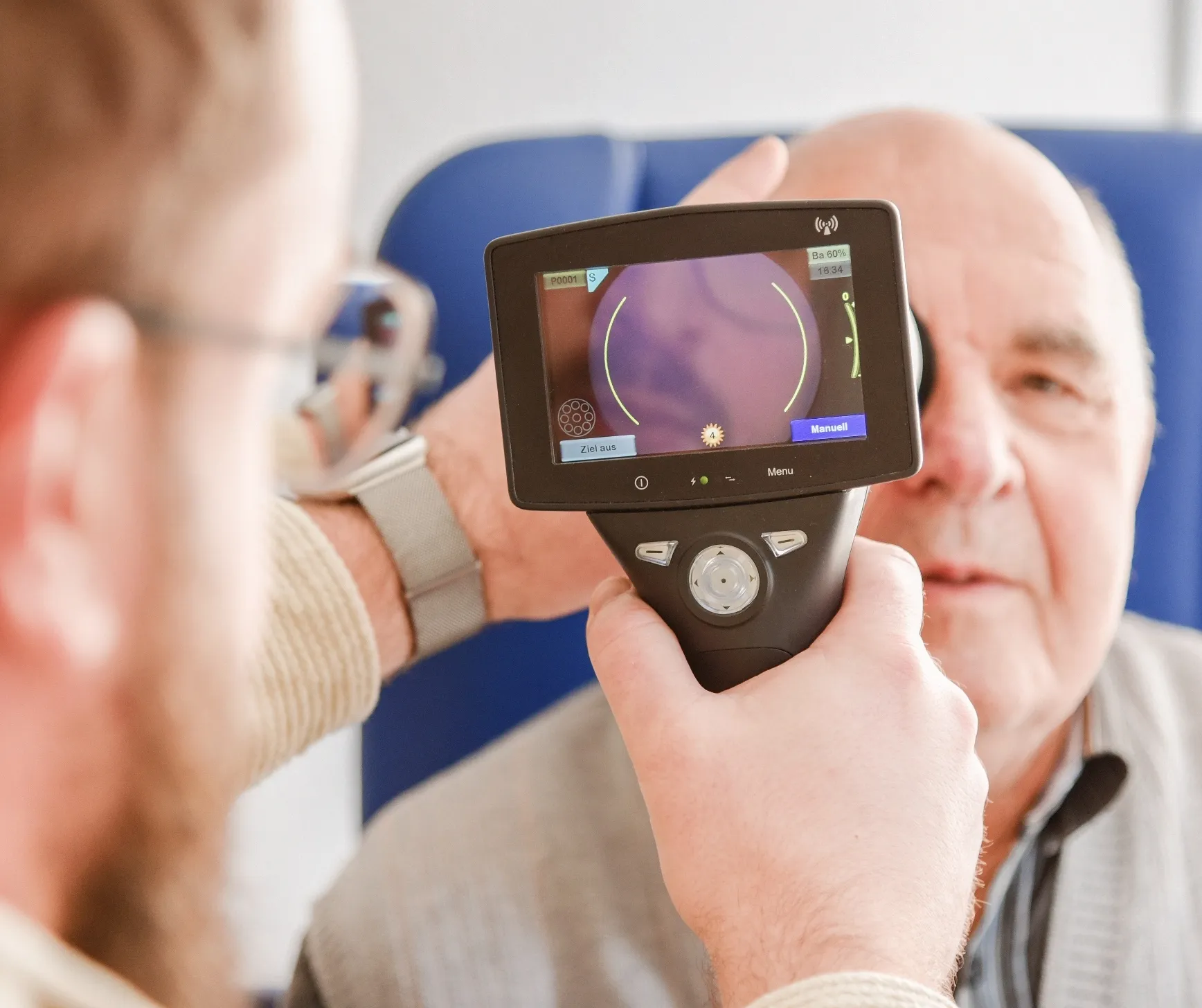 Ein Mann hält ein medizinisches Gerät mit Bildschirm vor das Auge eines älteren Mannes zur Augenuntersuchung.