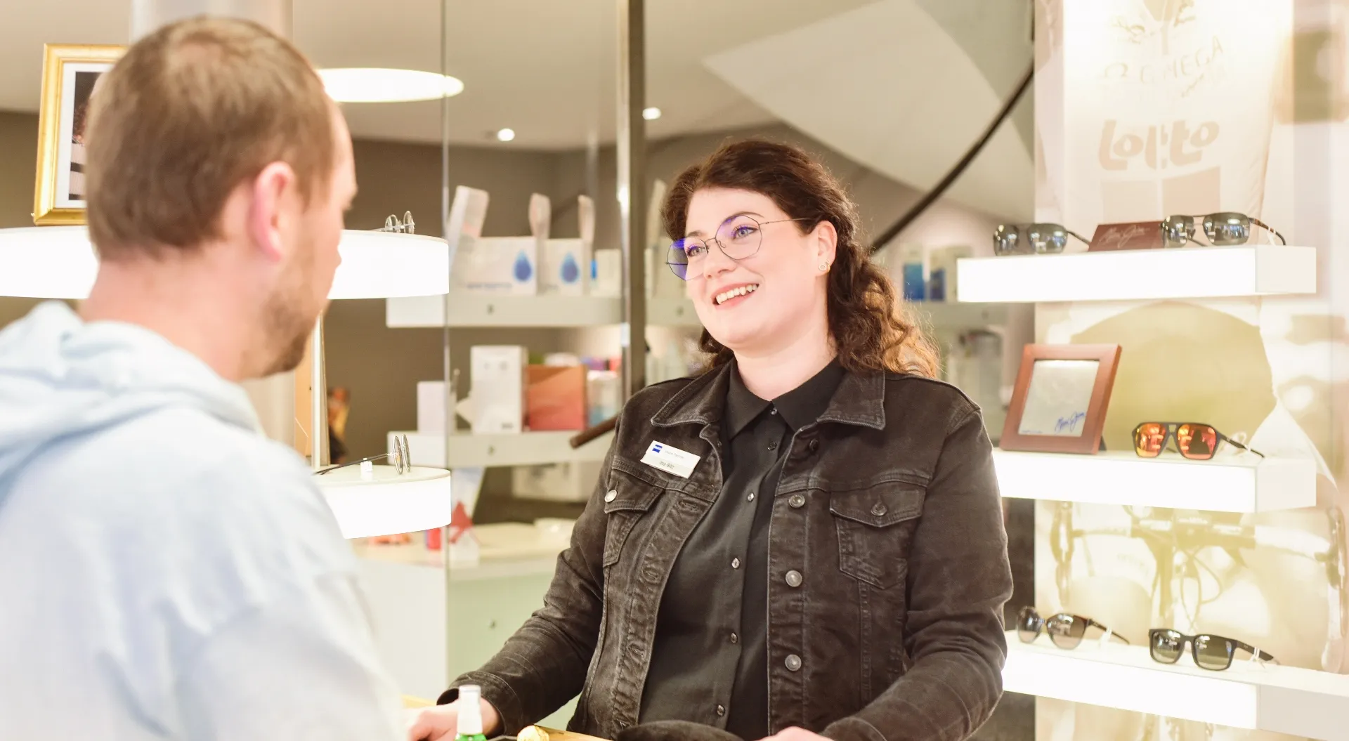 Lächelnde Verkäuferin mit Brille und schwarzer Jacke berät einen Kunden an einem Ladenlokal mit Brillenregalen im Hintergrund.