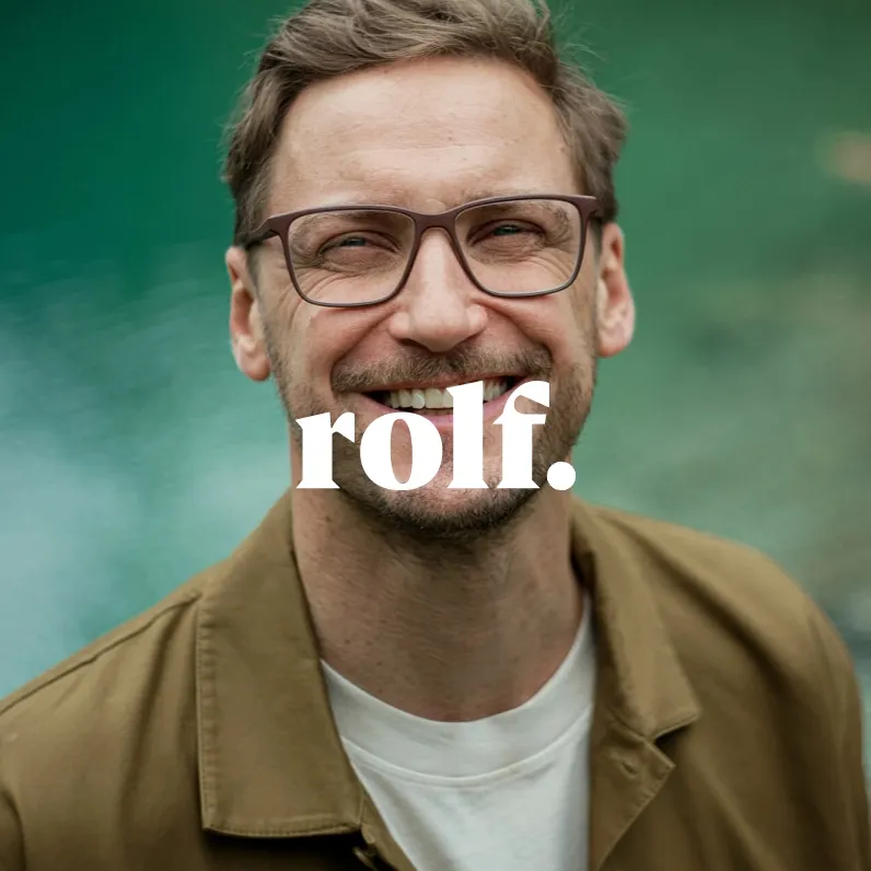Markenbild Rolf. Lächelnder Mann mit Brille und brauner Jacke vor unscharf grünem Hintergrund, Text ‚rolf.‘ in Weiß über dem Bild.