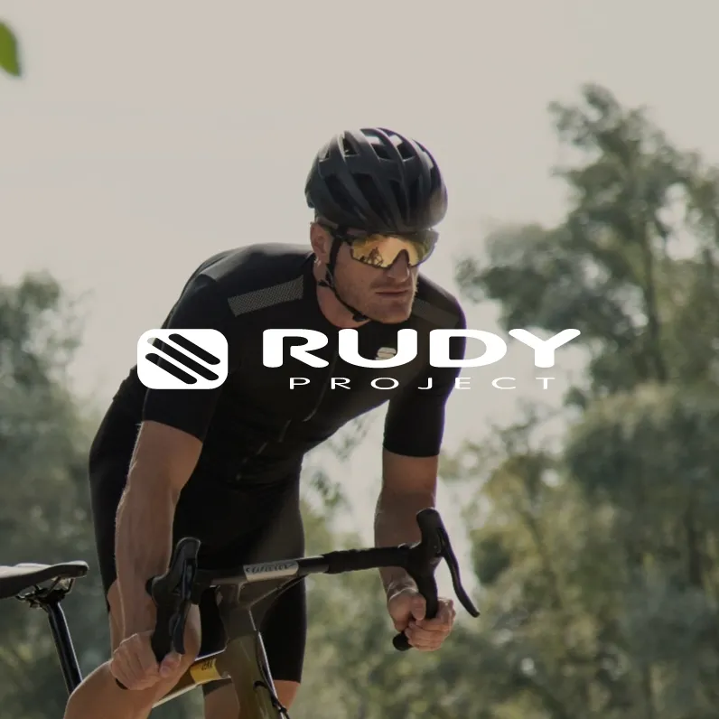 Markenbild Rudy Project. Radfahrer mit Helm und Sportbrille fährt Rennrad in natürlicher Umgebung mit Rudy Project Logo.
