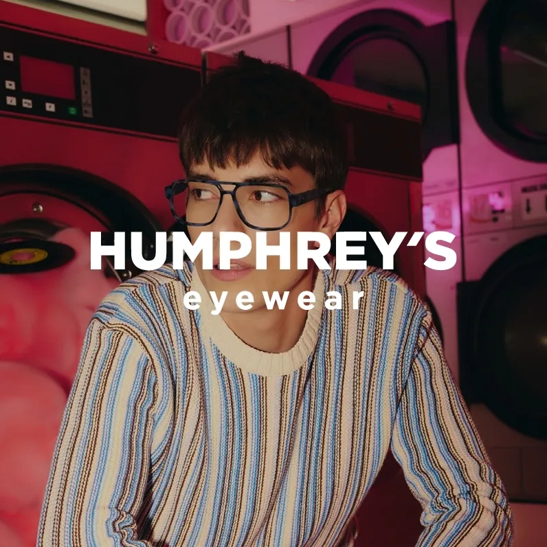 Markenbild Humphrey's eyewear. Junger Mann mit Brille und gestreiftem Pullover sitzt in einer Waschküche mit rosa Beleuchtung.