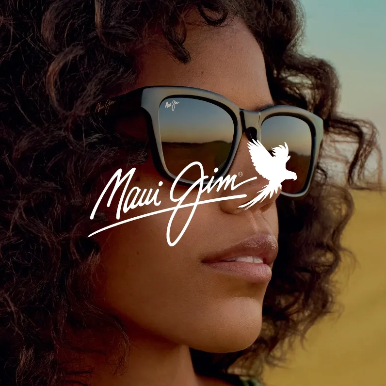 Markenbild Maui Jim. Nahaufnahme eines Gesichts mit lockigem Haar, die eine Sonnenbrille von Maui Jim trägt, in der sich ein Landschaftshorizont spiegelt.
